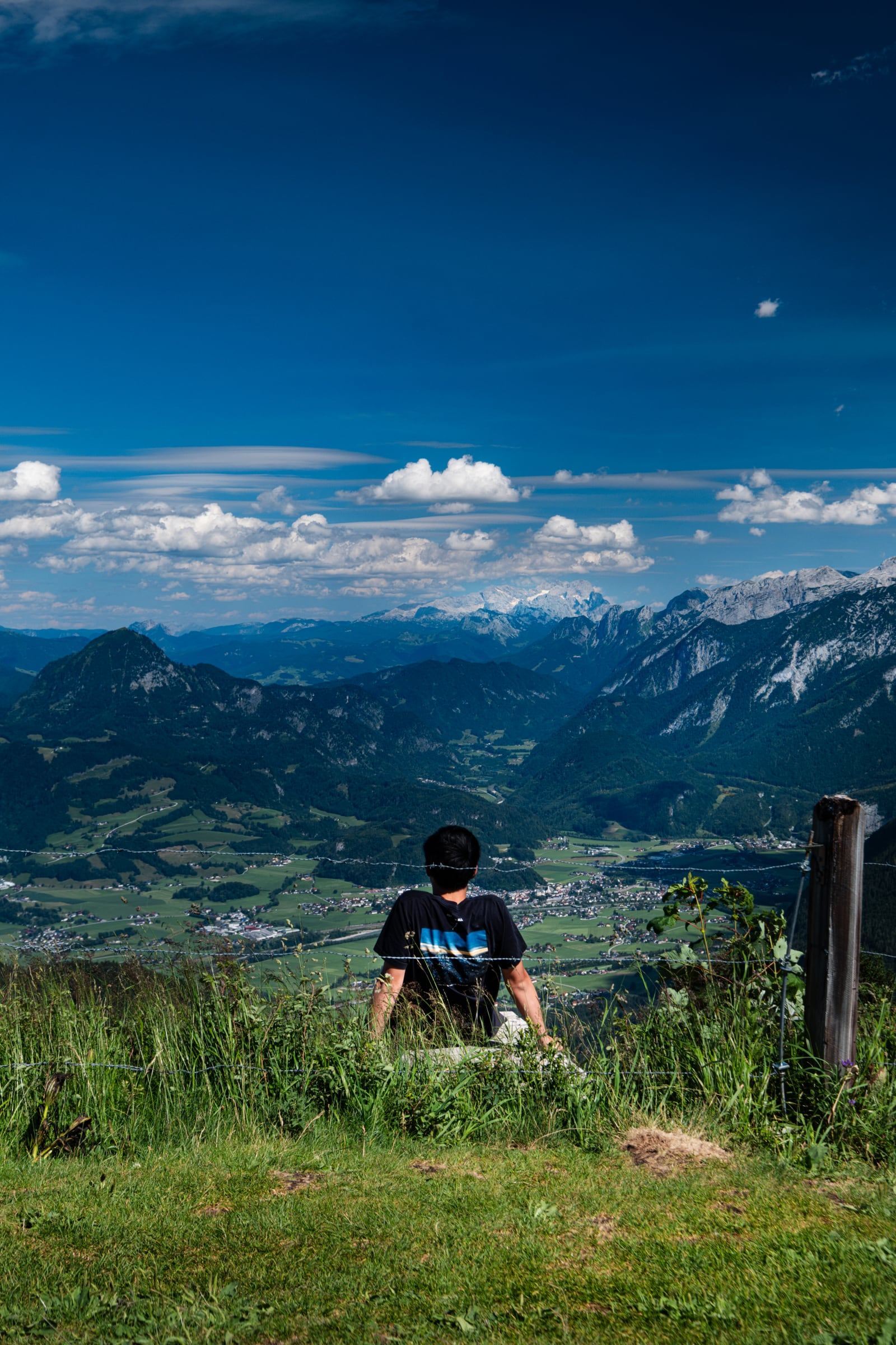 A man sits on a grassy summit looking out over a green valley toward snow-capped peaks.