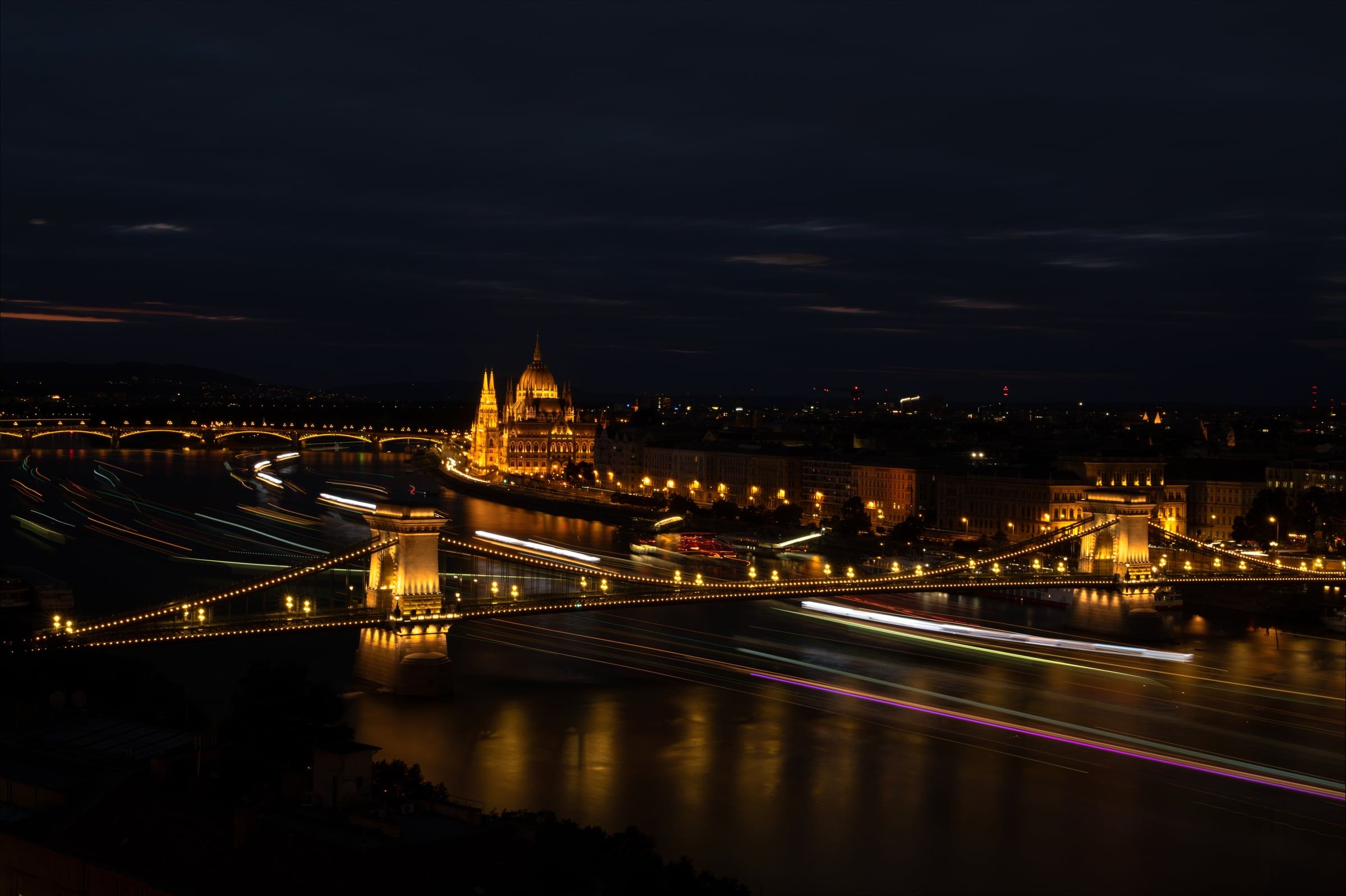 The illuminated Chain Bridge and Parliament across the Danube at night, with boat light trails on the river.