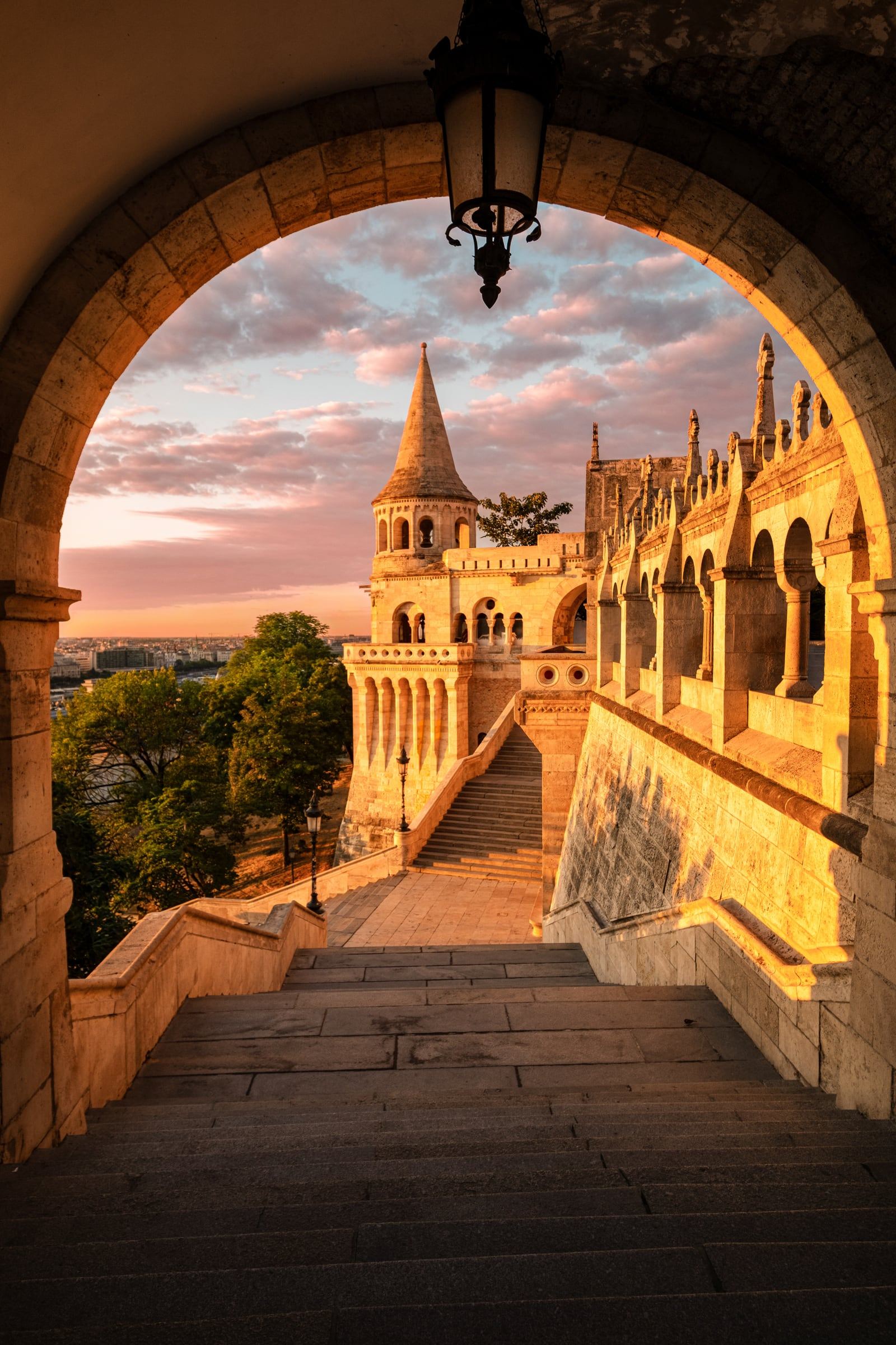 Stone steps lead through an arch toward the turrets of Fisherman's Bastion under a pink sky.