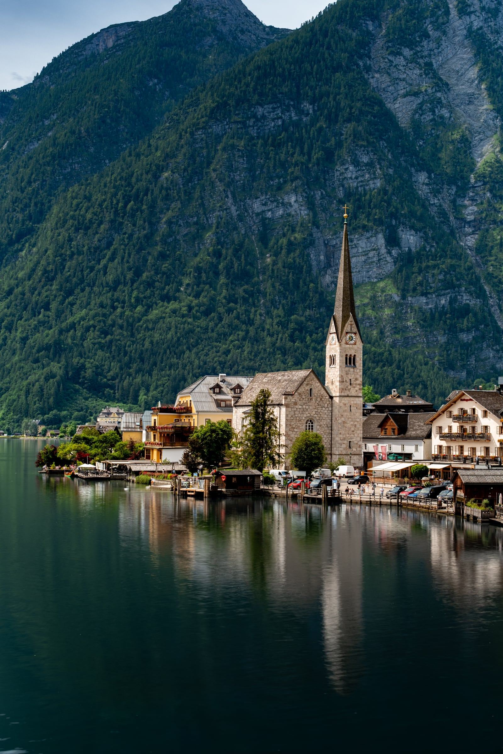 A church spire and lakeside houses reflected in still water beneath a steep forested mountain.