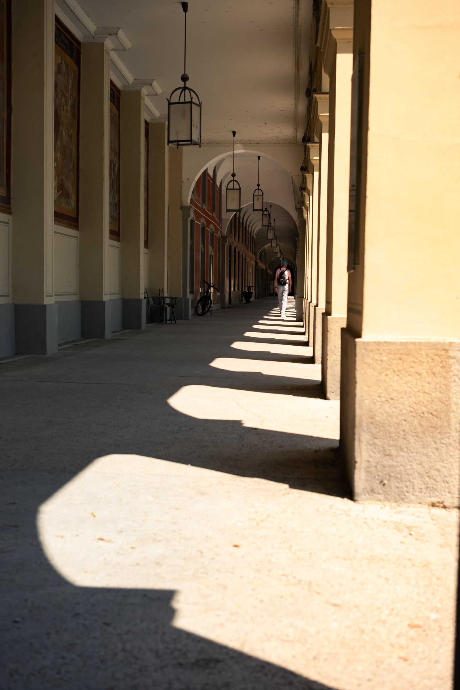 A figure walks down a long arcade where pillars cast bold shadows across the sunlit floor.