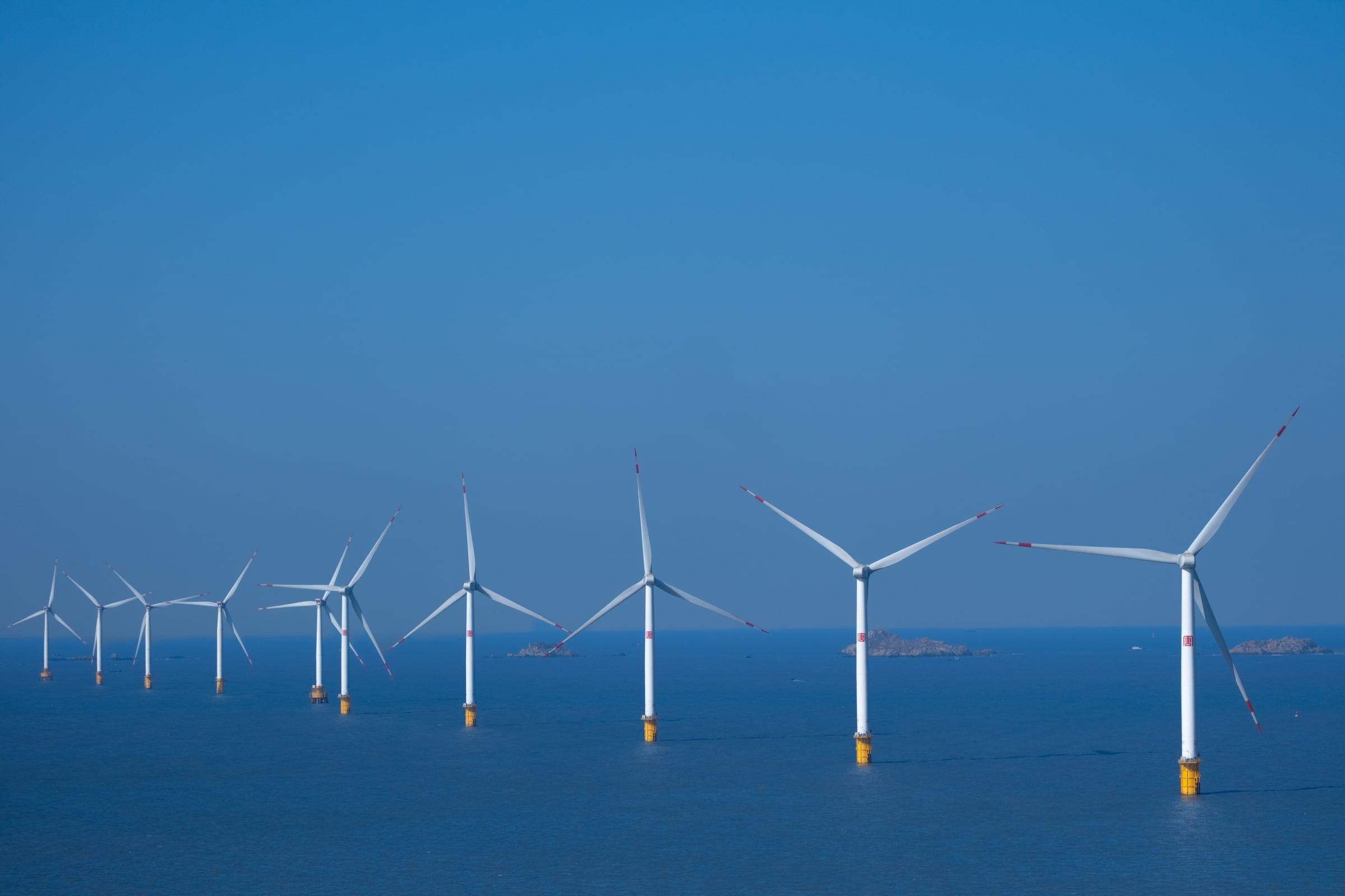 A row of white offshore wind turbines standing in a calm blue sea under a clear sky.