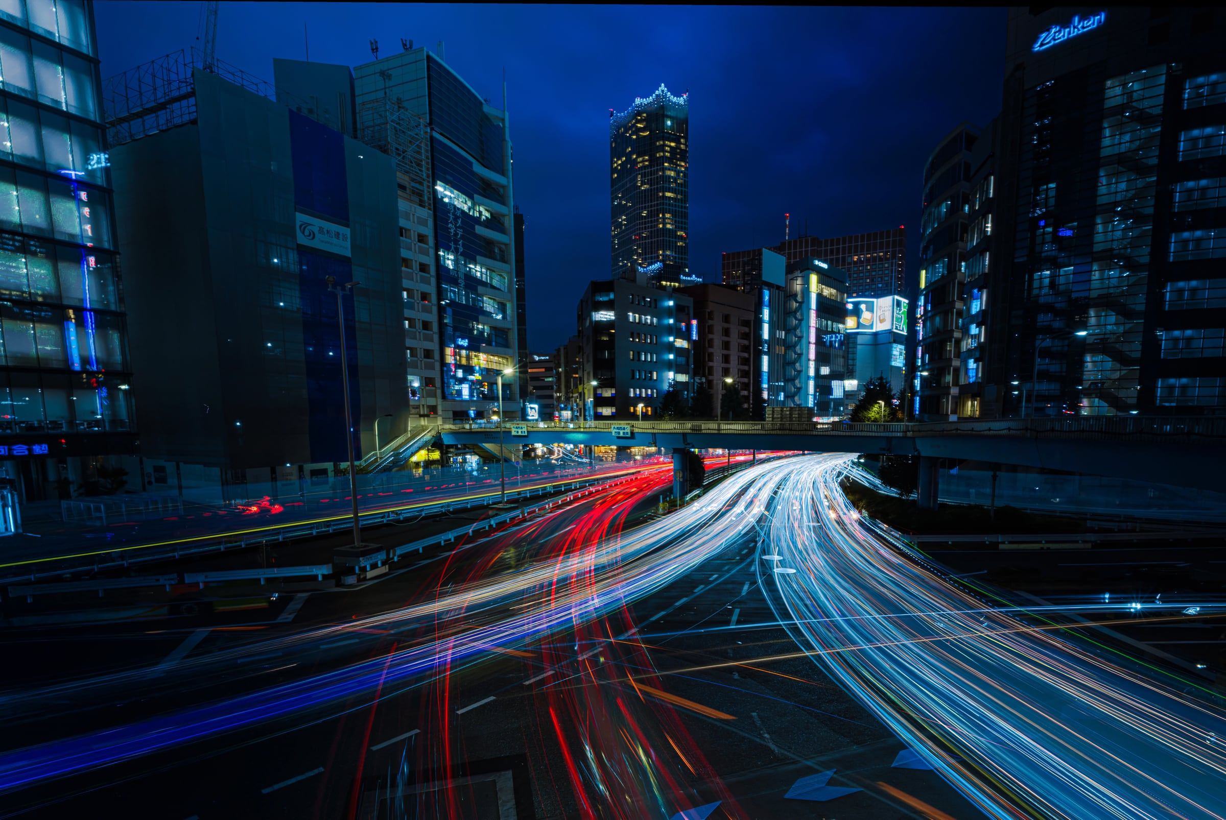 Long-exposure light trails sweep along a curving city expressway beneath lit towers at blue hour.